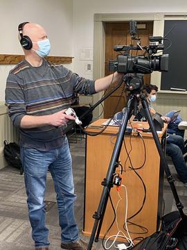 A videographer standing behind a professional camera on a portable tripod, holding a long metal arm to control the direction and zoom. The camera is attached with multiple cables to a laptop and to electrical power. In the background is a classroom door and chalkboard.